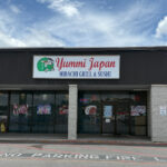 Front of Yummi Japan Hibachi Grill & Sushi, located in a shopping plaza with a white sign and red lettering above the entrance. Large glass windows display photos of menu items.