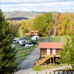 Brushcreek Falls RV Resort and Campground, featuring a two-story cabin-style building with stairs, several RVs parked along a winding road, and a backdrop of colorful fall foliage.