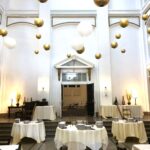 Interior of David's Downtown restaurant featuring tall white walls, round tables with white tablecloths, and gold and white decorations hanging from the ceiling.