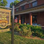 Baker's Hill Inn sign in front of a two-story red-brick inn with a covered porch, outdoor seating, and lush greenery in the front yard.