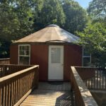 A round, red yurt with a white door and metal roof, accessed by a wooden deck surrounded by trees at Kairos Wilderness Resort.