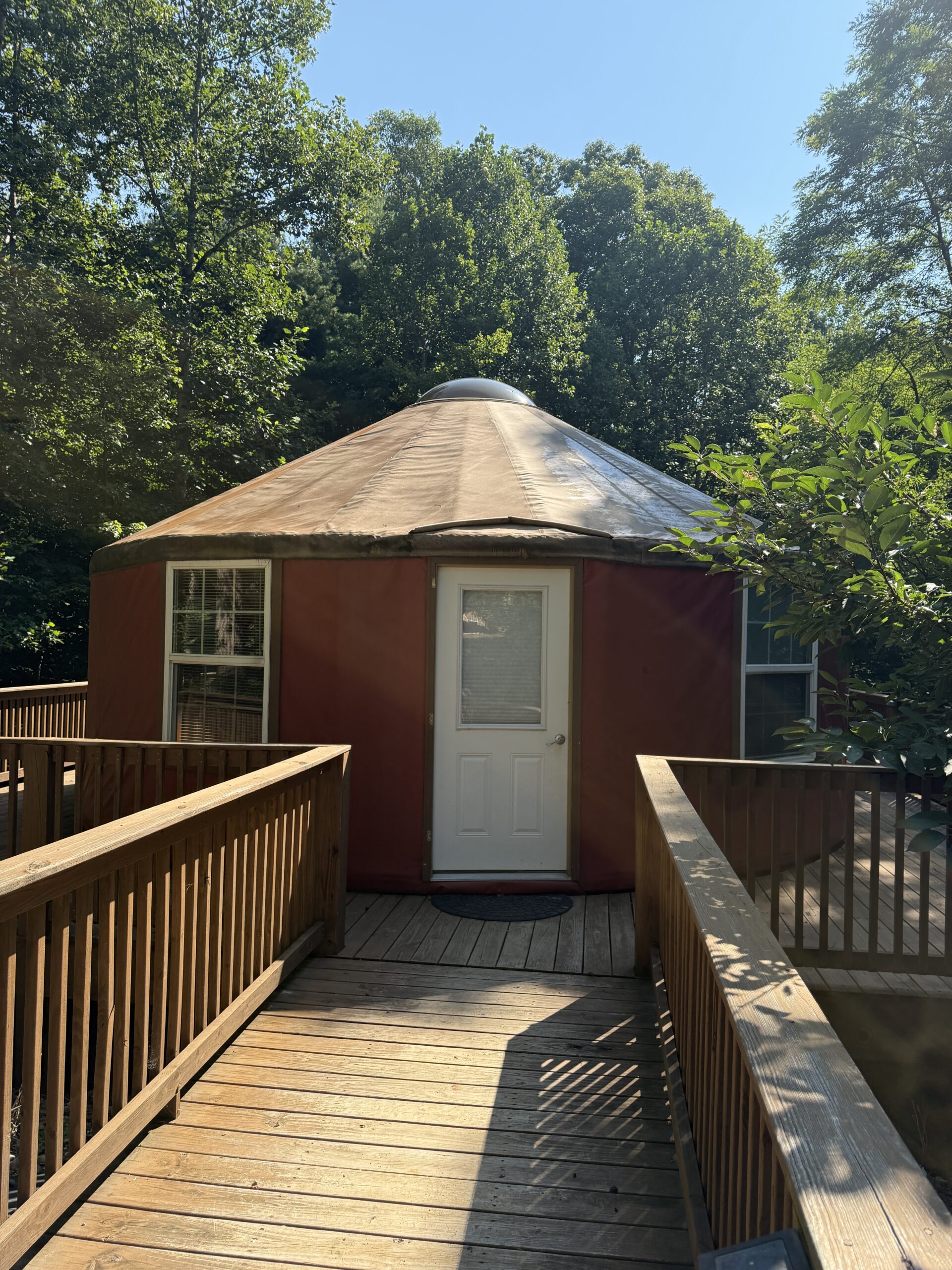 A round, red yurt with a white door and metal roof, accessed by a wooden deck surrounded by trees at Kairos Wilderness Resort.
