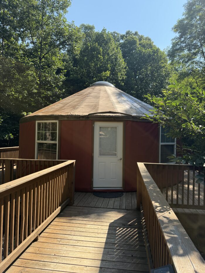 A round, red yurt with a white door and metal roof, accessed by a wooden deck surrounded by trees at Kairos Wilderness Resort.