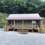 Single-story cabin with a metal roof and wooden porch railings at Gold and Blue Cabin Rentals, set against a wooded hillside with a circular stone fire pit in the gravel lot.