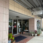 Entrance to The Ugly Duckling antique shop, featuring a sign that reads 'Antiques, Vintage Items, Old Books & More,' potted plants, and a small outdoor seating area along the sidewalk.