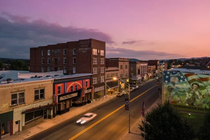 Downtown street at sunset with storefronts lit by string lights and a mural on a nearby building