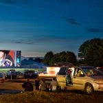 Cars parked at Pipestem Drive-In at sunset, with people sitting on the ground and a movie playing on the outdoor screen.