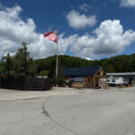Appalachian RV Resort and Campground entrance with a log-style check-in building, large American flag, and RVs in the background under a partly cloudy sky.