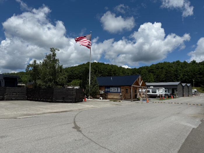 Appalachian RV Resort and Campground entrance with a log-style check-in building, large American flag, and RVs in the background under a partly cloudy sky.