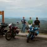 Three motorcycles parked at the East River Mountain Overlook in Mercer County, West Virginia, as three riders stand facing the mountain view beside the Almost Heaven swing at sunset.