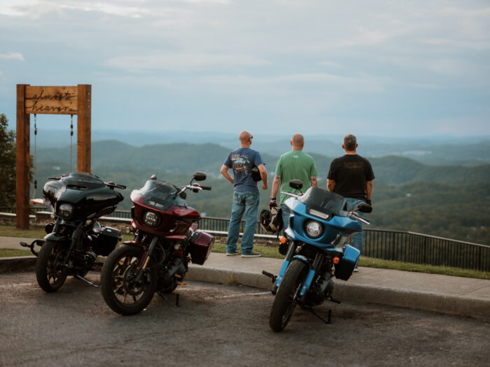 Three motorcycles parked at the East River Mountain Overlook in Mercer County, West Virginia, as three riders stand facing the mountain view beside the Almost Heaven swing at sunset.