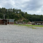ATV Trail Camp with multiple small cabin-style buildings arranged in a row, set on a large gravel lot surrounded by wooded hills.