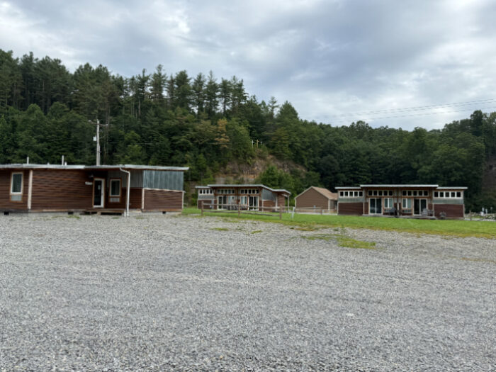 ATV Trail Camp with multiple small cabin-style buildings arranged in a row, set on a large gravel lot surrounded by wooded hills.