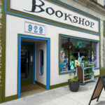 Front of Hatters Bookshop, featuring a blue-trimmed entrance, large illustrated display window, floral mural accents, and a sidewalk chalkboard sign. A planter and small book cart sit outside the window.