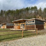 Modern cabins with wood and corrugated metal siding sit along a gravel drive in a wooded Appalachian setting, with tall pine trees and rocky hillsides rising behind the lodging area under a partly cloudy sky.