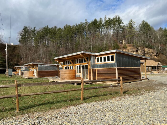 Modern cabins with wood and corrugated metal siding sit along a gravel drive in a wooded Appalachian setting, with tall pine trees and rocky hillsides rising behind the lodging area under a partly cloudy sky.