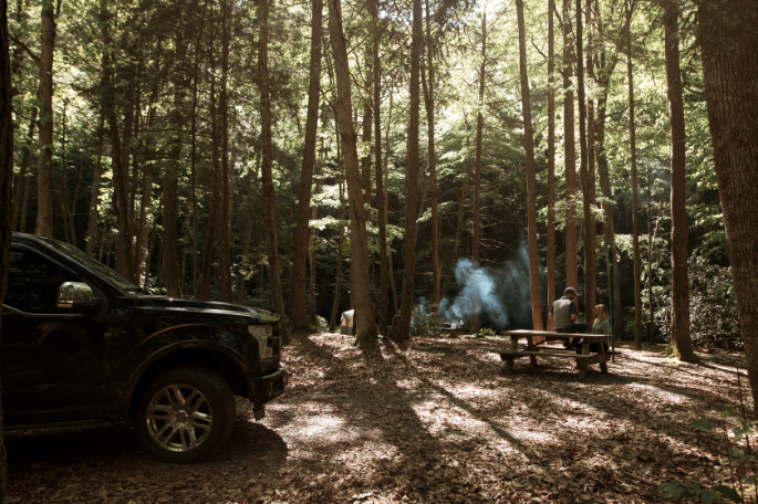 Image of Camp Creek, showing a shaded wooded campsite with a black pickup truck parked on the left, a picnic table with two people seated on the right, and smoke rising from a campfire in the background.