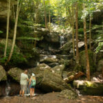 Image of White Oak Waterfall, showing two people standing at the base surrounded by large moss-covered rocks, trees, and a cascading waterfall in the background.