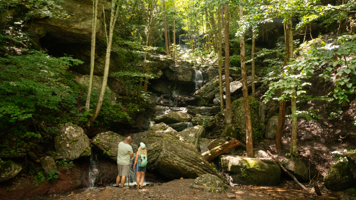 Image of White Oak Waterfall, showing two people standing at the base surrounded by large moss-covered rocks, trees, and a cascading waterfall in the background.