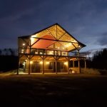 Lake Mountain Manor at dusk, a large timber-frame lodge glowing with warm lights, featuring high peaked roof, wrap around balcony, and open structural beams.