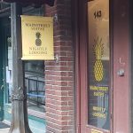 Entrance to Main Street Suites with a brick exterior, large pineapple graphic on the door, and a yellow hanging sign that reads 'Main Street Suites Nightly Lodging.'"