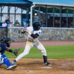 Ridge Runners Baseball player at bat during a game, with a catcher crouched behind the home plate and spectators watching from the stands.
