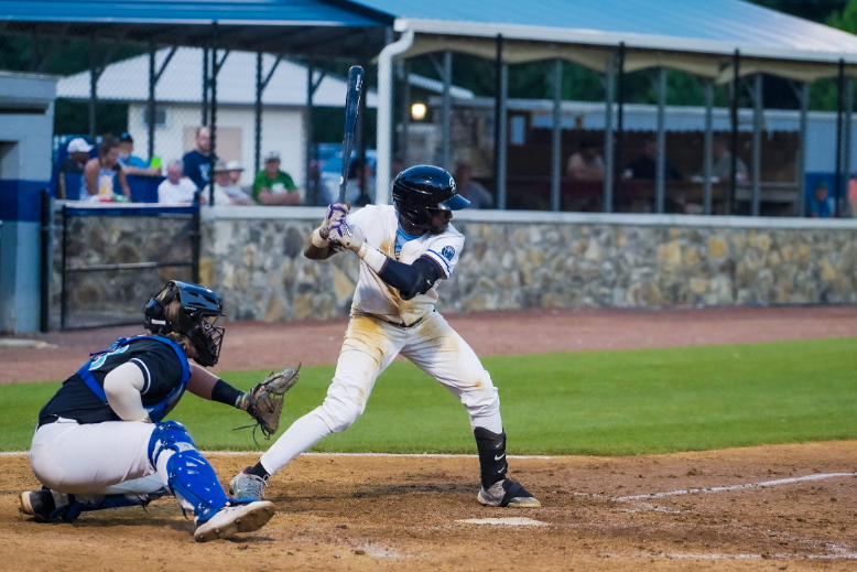 Ridge Runners Baseball player at bat during a game, with a catcher crouched behind the home plate and spectators watching from the stands.