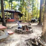 Rustic wooden cabin at Pierce's Place nestled in the woods, with picnic tables, a fire pit surrounded by stones, and an ATV parked nearby.