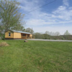 Wooden cabin with a covered porch at Hidden Hollow ATV Ranch, set on a grassy hilltop with trees and mountains under a sunny sky.