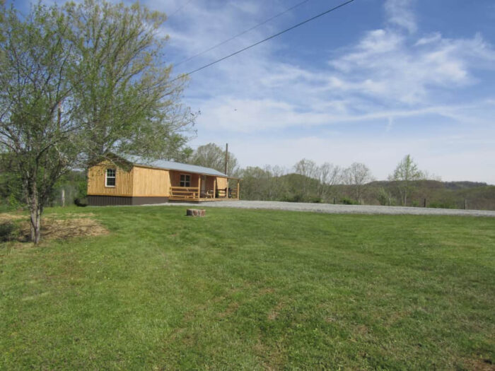 Wooden cabin with a covered porch at Hidden Hollow ATV Ranch, set on a grassy hilltop with trees and mountains under a sunny sky.