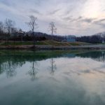 Lakeview Campground with still water reflecting bare trees and a cloudy sky, surrounded by hills and scattered buildings.