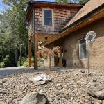 Exterior view of On Cue Properties LLC building, featuring wood and stone textures, a gravel yard with decorative landscaping, and surrounded by trees.