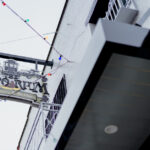 Hanging sign for The Emporium: Adventures & Hobbies, mounted on a white building with colorful string lights and a black overhang visible above.