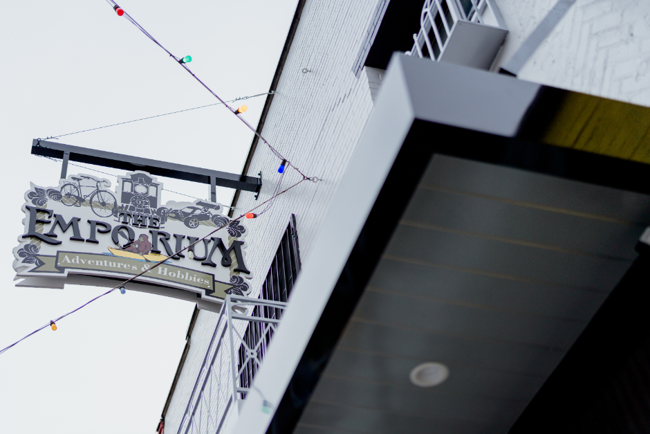 Hanging sign for The Emporium: Adventures & Hobbies, mounted on a white building with colorful string lights and a black overhang visible above.