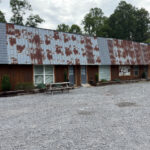 Exterior of Pinnacle Place ATV Lodge, a rustic type building with a long, weathered metal roof, wood-paneled walls, multiple windows, and picnic tables in a gravel lot.