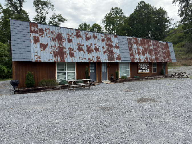 Exterior of Pinnacle Place ATV Lodge, a rustic type building with a long, weathered metal roof, wood-paneled walls, multiple windows, and picnic tables in a gravel lot.