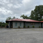Single-story Rest & Ride ATV Lodge with a red metal roof, light gray exterior, covered entrance, and shrubs lining the front.