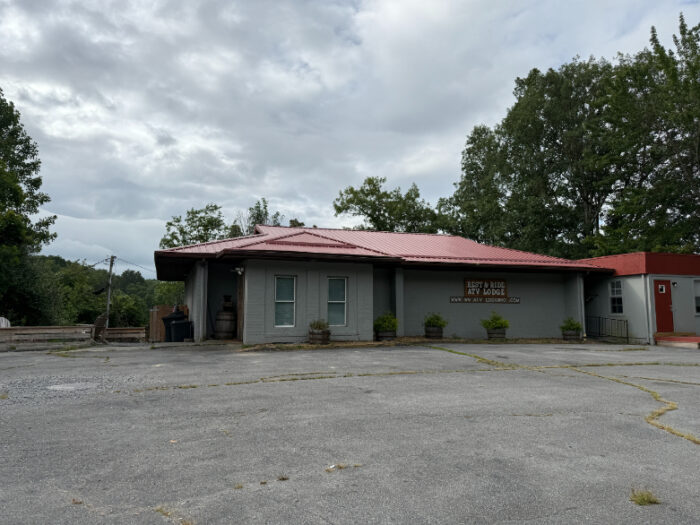 Single-story Rest & Ride ATV Lodge with a red metal roof, light gray exterior, covered entrance, and shrubs lining the front.