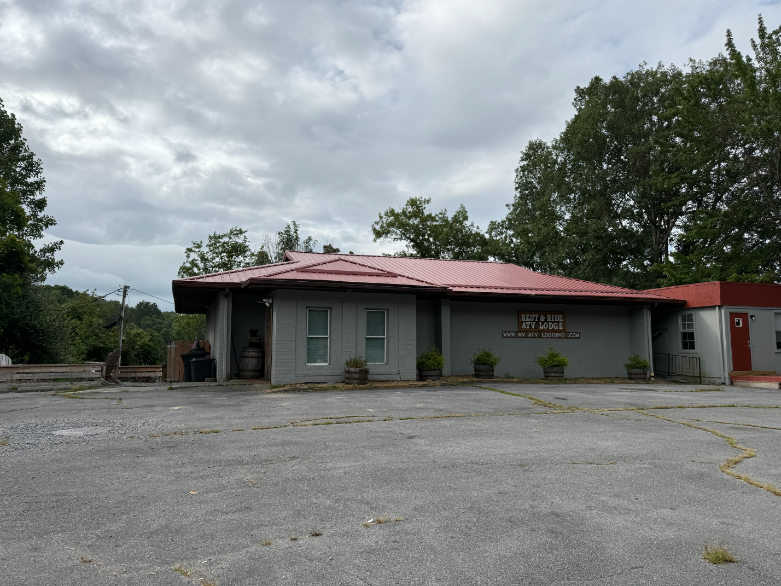 Single-story Rest & Ride ATV Lodge with a red metal roof, light gray exterior, covered entrance, and shrubs lining the front.
