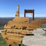 Wooden piece shaped like the state of West Virginia with the words 'almost heaven' on it, positioned in front of a scenic swing overlooking a mountain view.