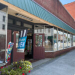 Wearhouse Clothing storefront with a green awning, large glass display windows, and maroon double doors. A blue 'We Are OPEN' banner and colorful flags are displayed near the entrance, with potted flowers on the sidewalk.