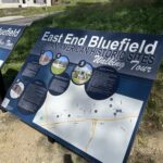 Informational sign for East End Walking Trail Tour, featuring a map and photos of notable locations, mounted outdoors in a grassy area.