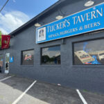 Exterior of Tucker's Tavern, a dark gray building with a blue sign that reads "Wings, Burgers, and Beers." The front features large windows with beer and sports decals, a red flag, and a small entrance near the parking lot.