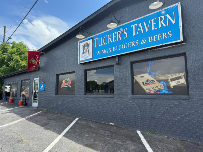 Exterior of Tucker's Tavern, a dark gray building with a blue sign that reads "Wings, Burgers, and Beers." The front features large windows with beer and sports decals, a red flag, and a small entrance near the parking lot.