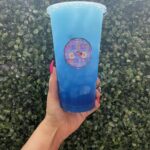 A vibrant blue loaded tea from Pure Bliss Nutrition, held up in front of a leafy green background. The clear plastic cup features the shop's colorful logo.