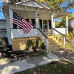 The Farmhouse at More Acres, a white cottage-style home with a front porch, American flag, and yellow steps leading to a cozy entryway. Surrounded by small shrubs and trees under a clear blue sky.