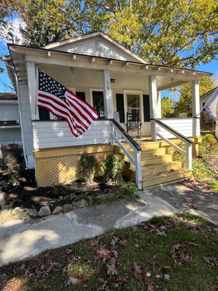 The Farmhouse at More Acres, a white cottage-style home with a front porch, American flag, and yellow steps leading to a cozy entryway. Surrounded by small shrubs and trees under a clear blue sky.