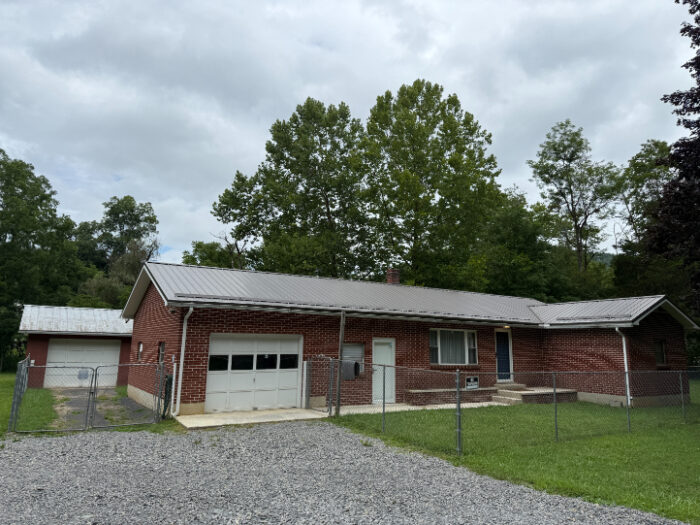 Single-story red brick house called Pappy's Trail House, featuring a metal roof, attached garage, gravel driveway, and fenced front yard with trees.