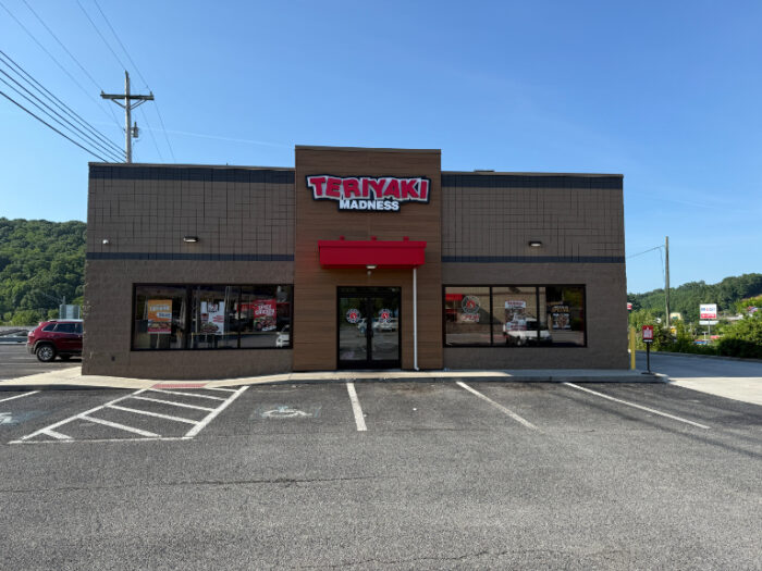 Front view of Teriyaki Madness restaurant with a red awning, brown and tan exterior, and large windows displaying promotional posters.