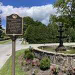 Historical marker sign and stone fountain surrounded by flowers at the start of the Bluefield Historical Home Driving Tour, with trees and a blue sky in the background.